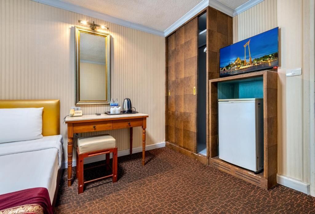 A hotel room with a bed, a desk with a telephone, a mirror, a refrigerator, and a painting on the wall.