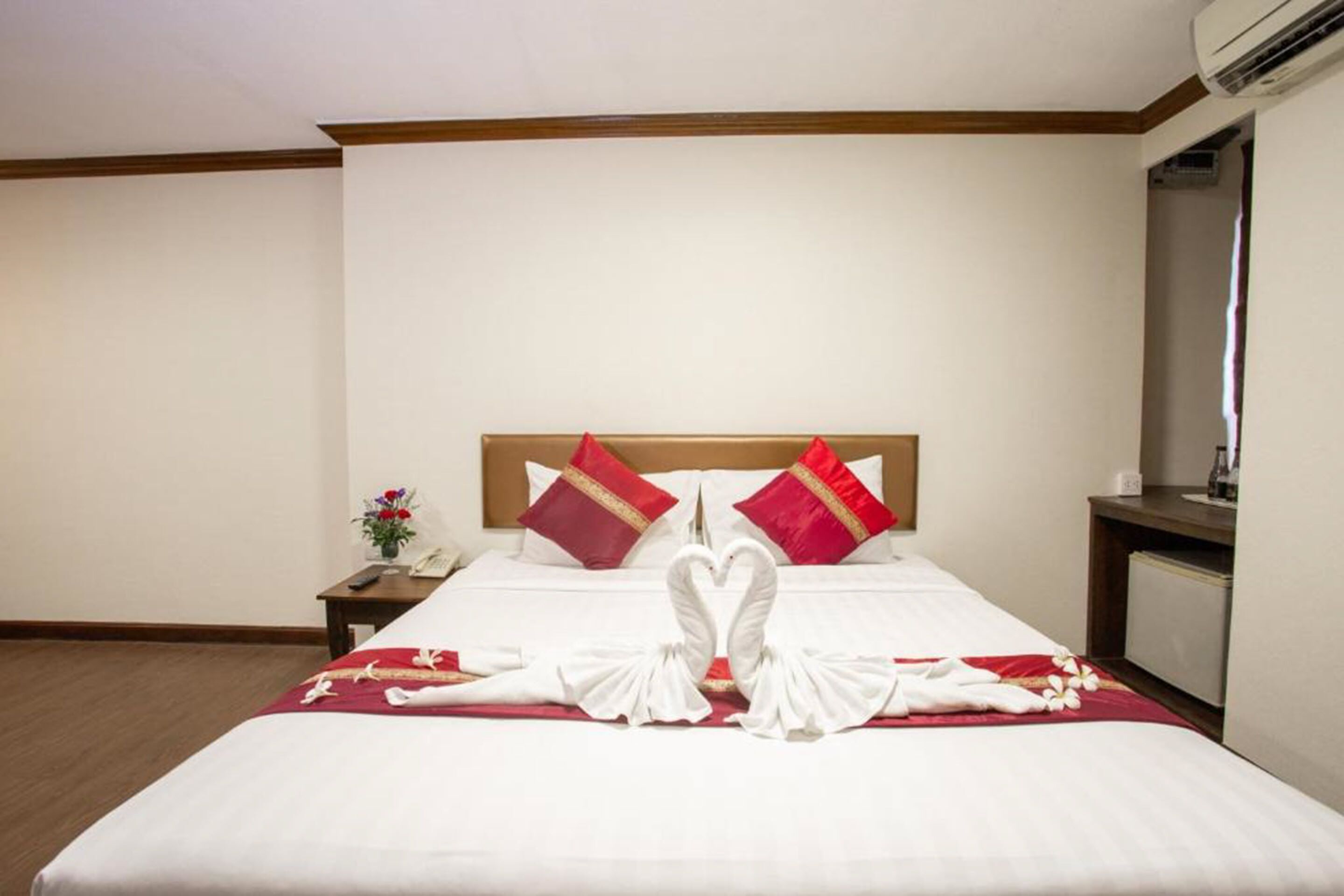 Une chambre d’hôtel avec un lit parfaitement fait, deux oreillers rouges, une décoration en forme de cœur réalisée avec des serviettes et une petite table de chevet sur laquelle est posé un vase de fleurs.