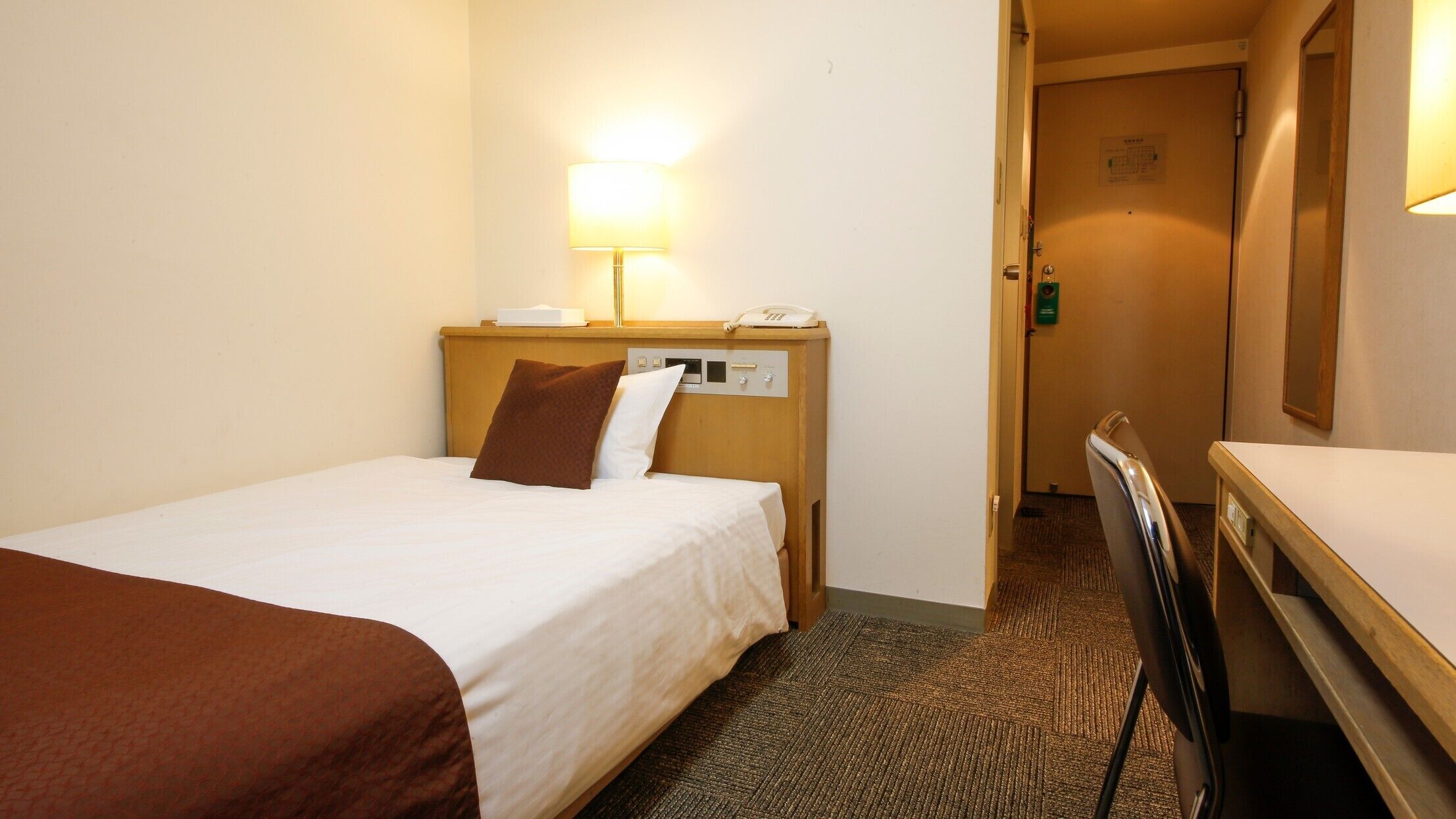 Hotellihuone, jossa on sänky, yöpöytä lampun kanssa, puhelin ja ovi, joka johtaa toiseen huoneeseen.