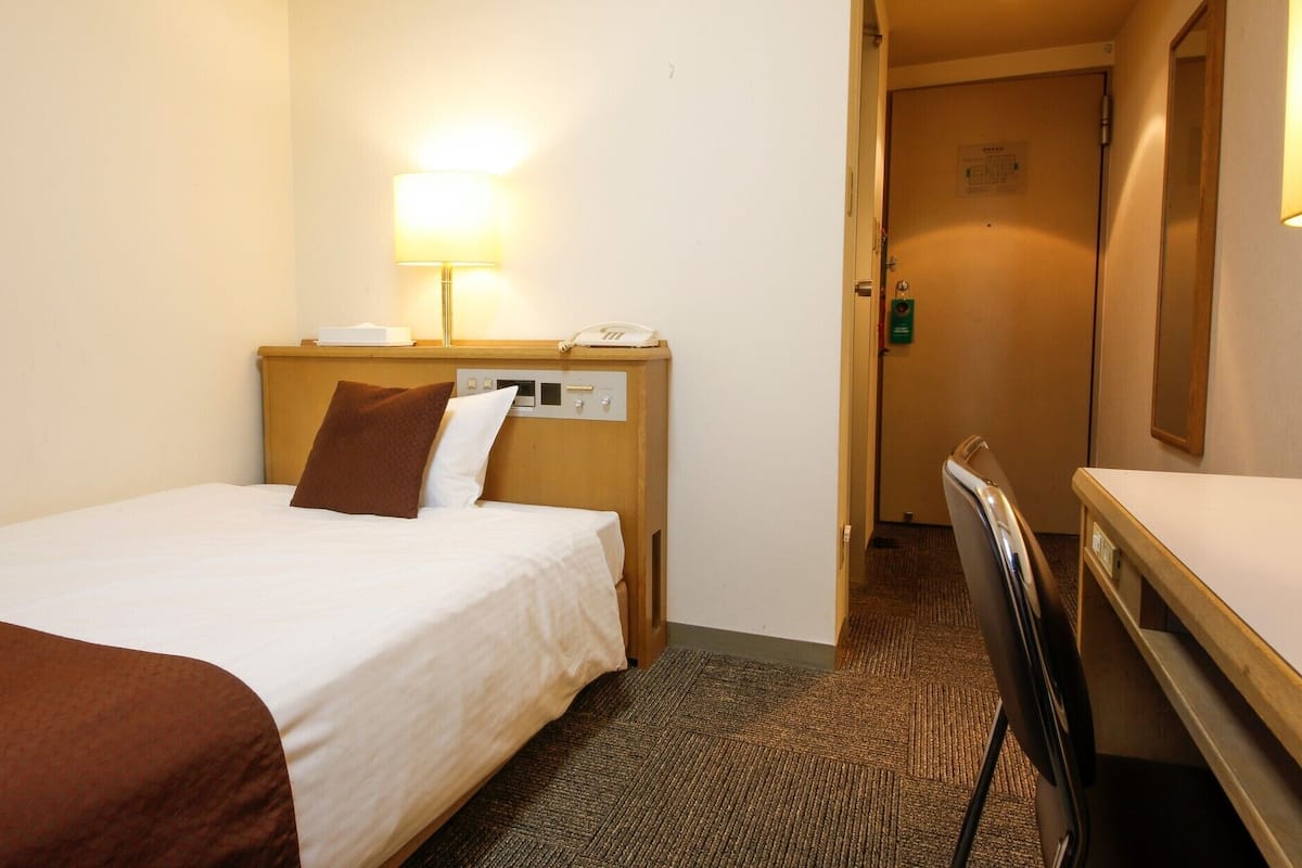 Hotellihuone, jossa on sänky, yöpöytä lampun kanssa, puhelin ja ovi, joka johtaa toiseen huoneeseen.