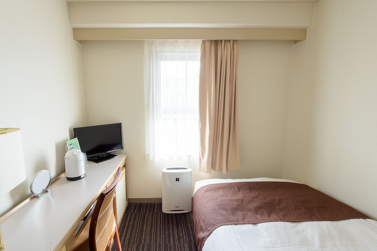 Ein Hotelzimmer mit Bett, Schreibtisch mit Computer und Kaffeemaschine, Fenster mit Vorhängen sowie einer Wandlampe.