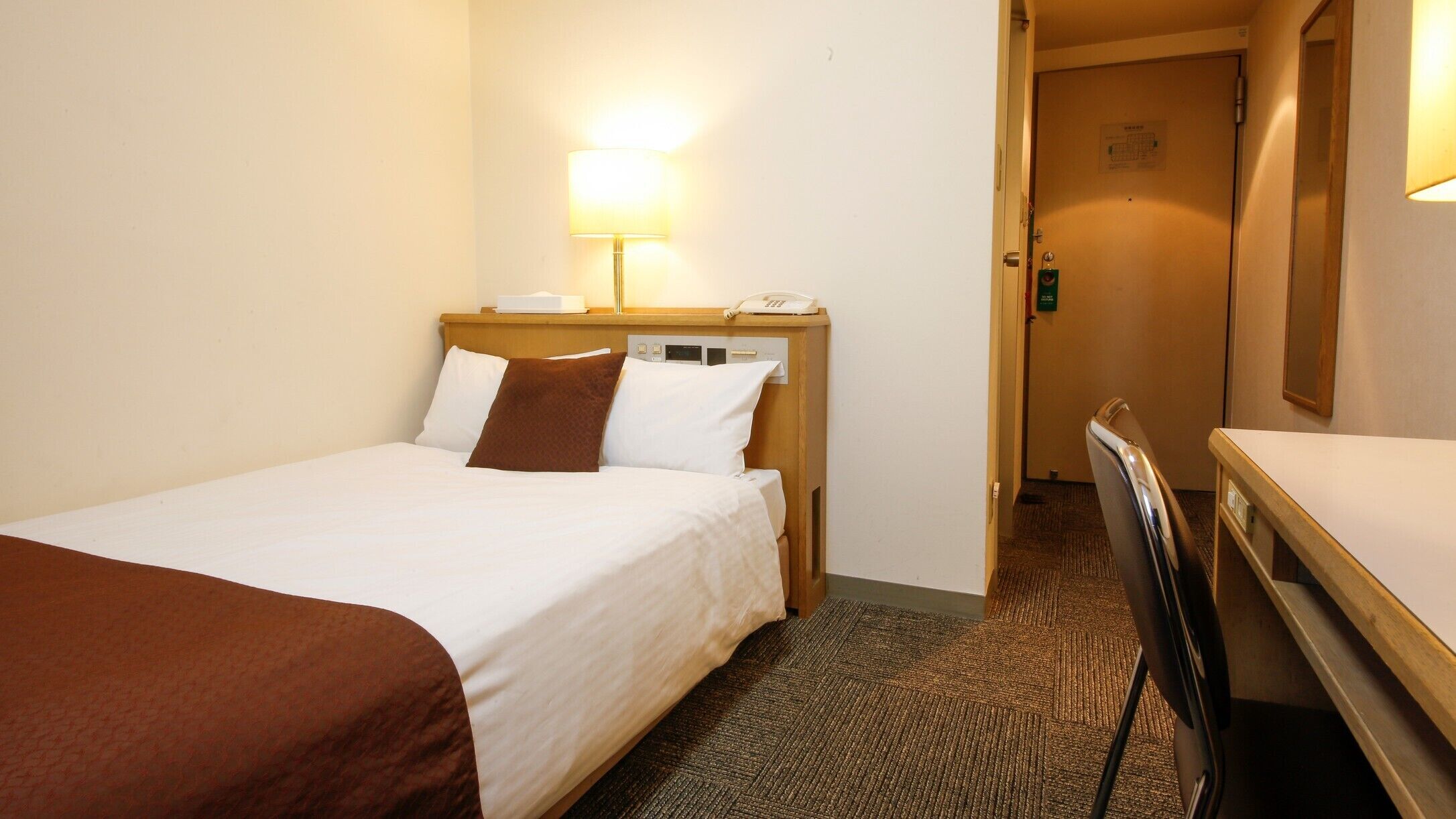 Hotellihuone, jossa on sänky, yöpöytälamppu, puhelin ja työpöytä tuolilla.