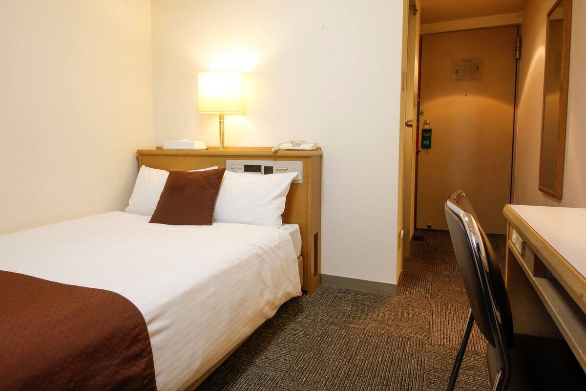Ein Hotelzimmer mit Bett, Nachttischlampe, Telefon und Schreibtisch mit Stuhl.