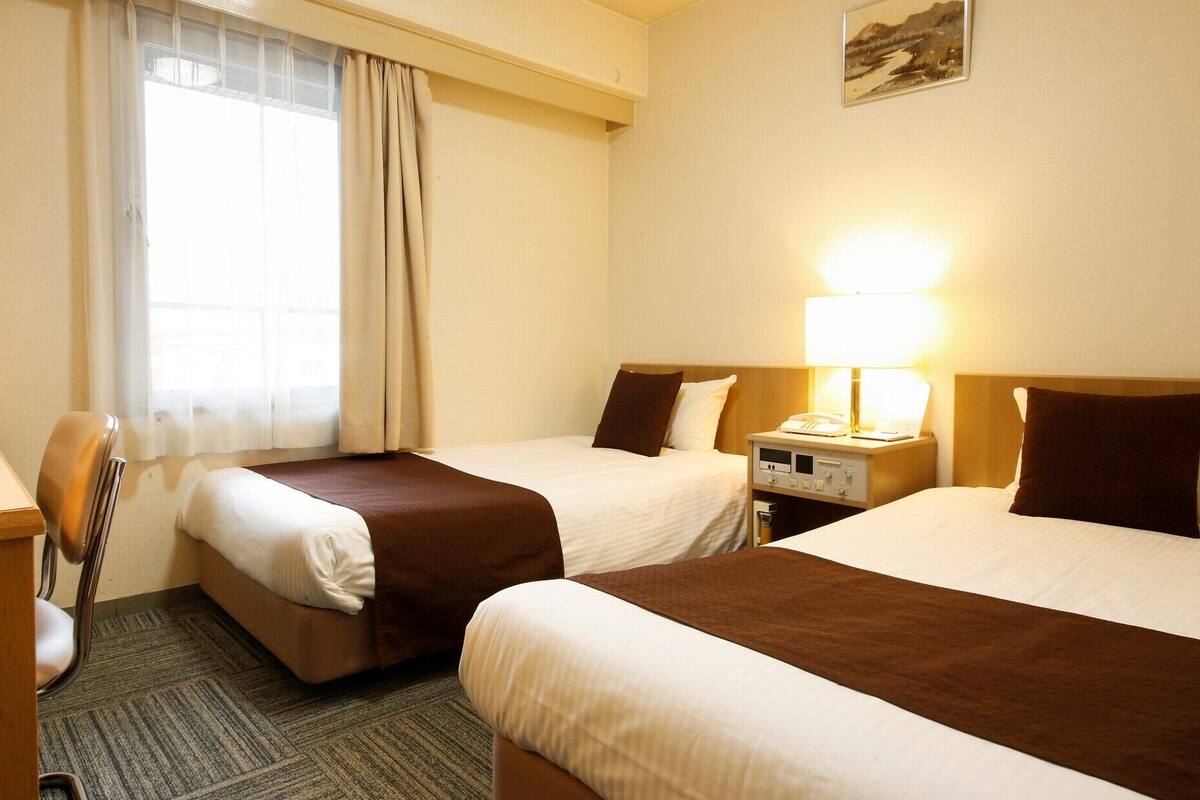 Ein Hotelzimmer mit zwei Betten, einem Schreibtisch, einem Stuhl, einem Fernseher und einem Fenster mit Vorhängen.