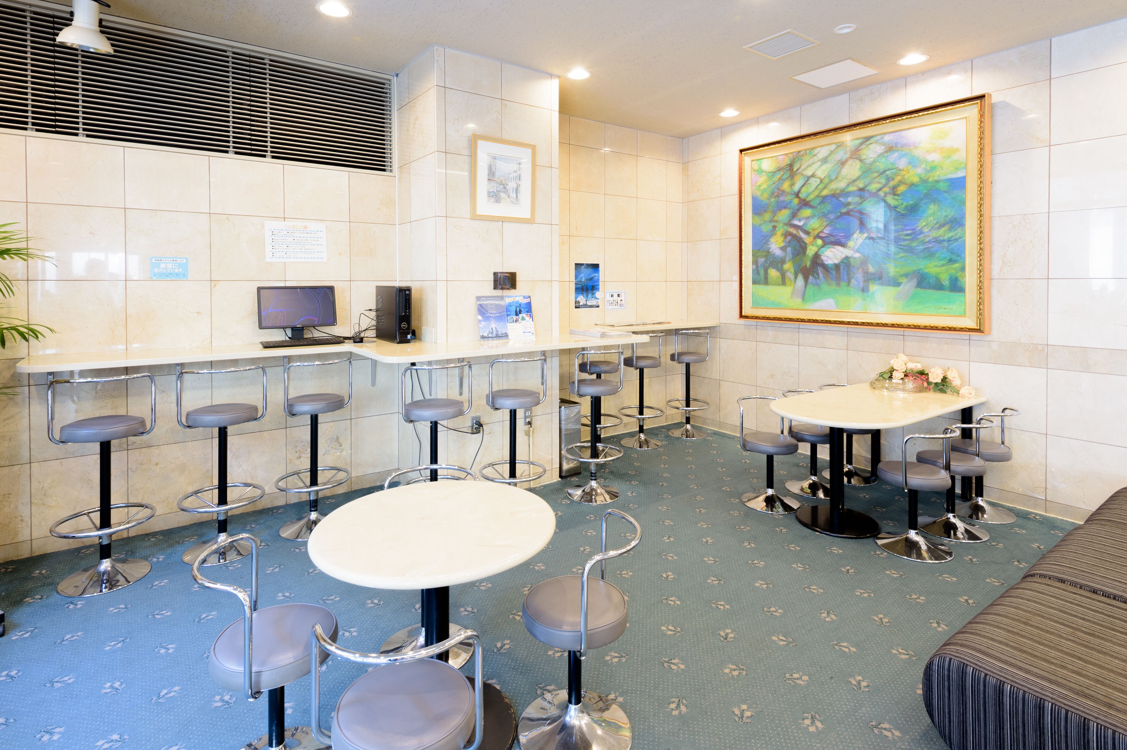 A hotel lobby with a reception desk, multiple bar stools, a round table, and a large painting on the wall.