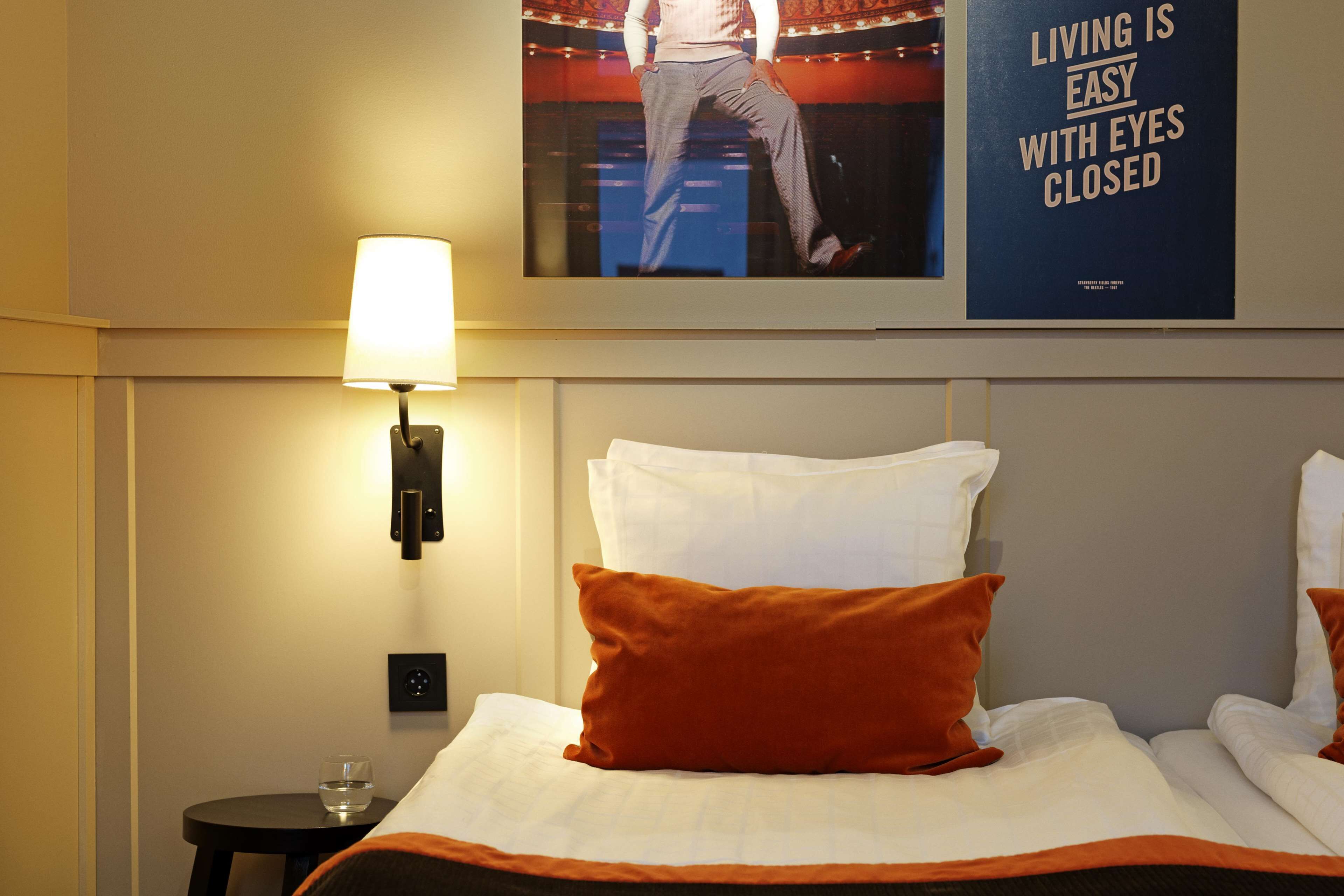 A hotel room with a bed, pillows, a lamp, a poster, and a small table with a glass of water.