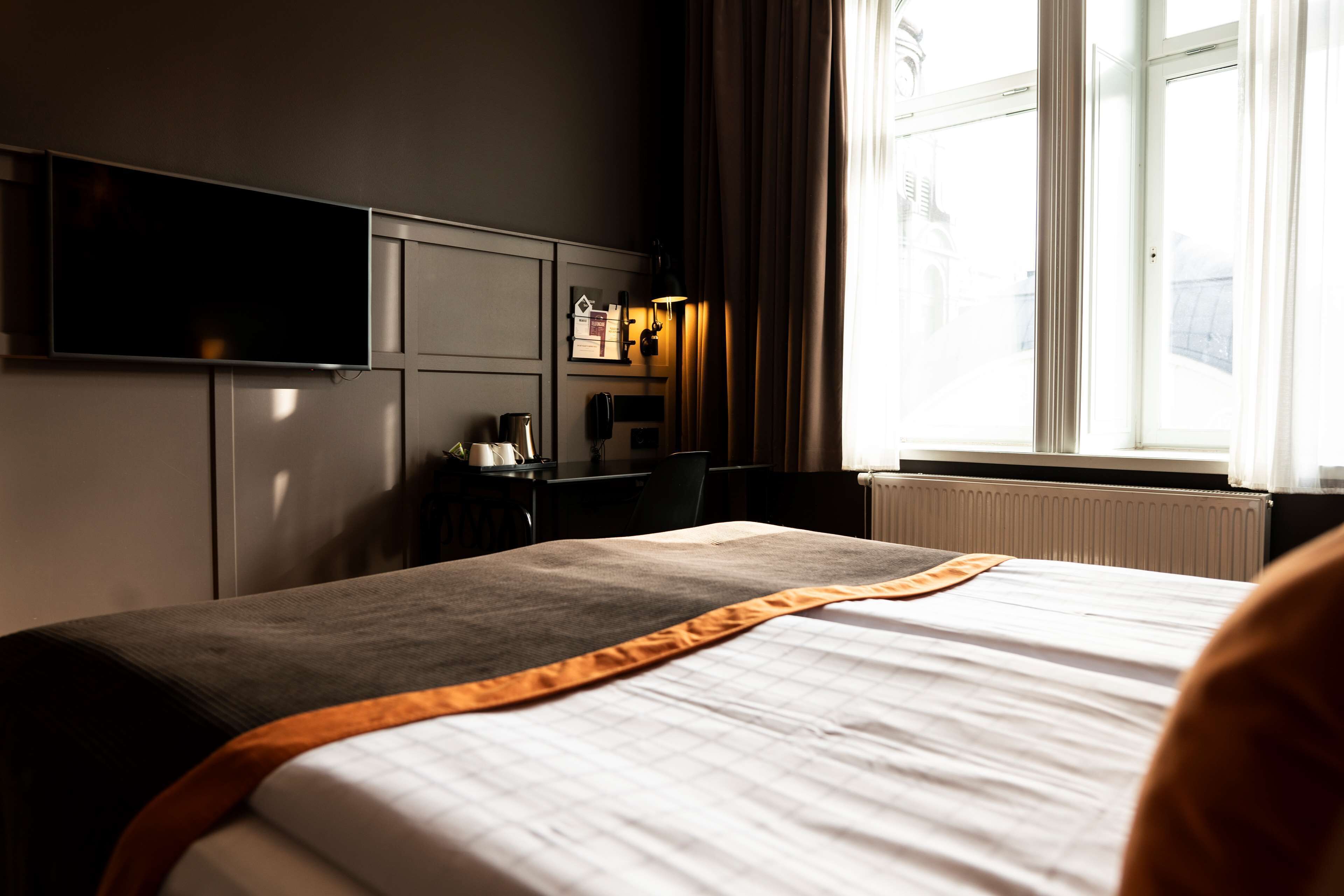 A hotel room with a large bed, a flat-screen TV mounted on the wall, a desk with a lamp, and a window with curtains.
