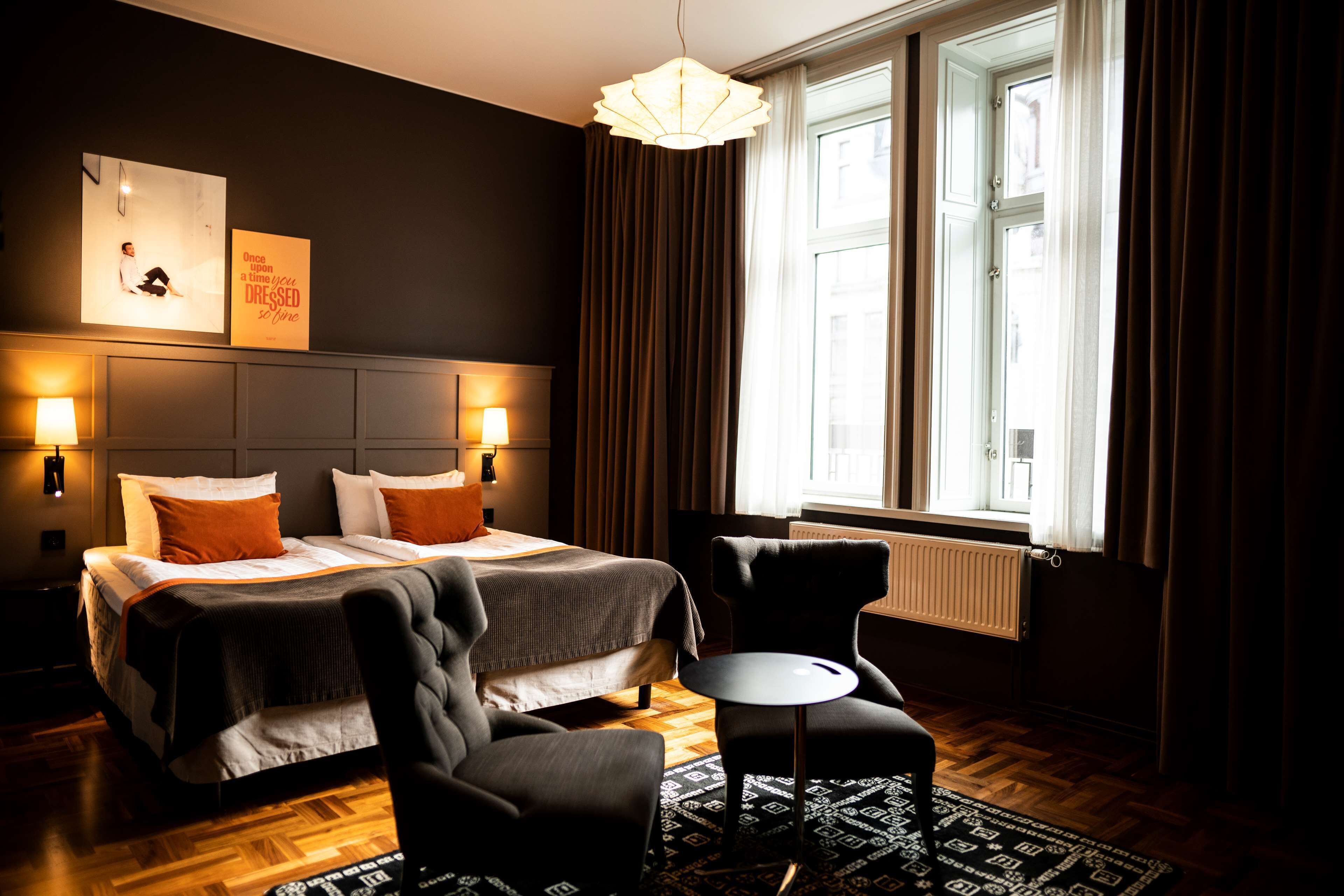A hotel room with a large bed, two bedside lamps, a chair, a small table, a patterned rug, and a window with curtains.