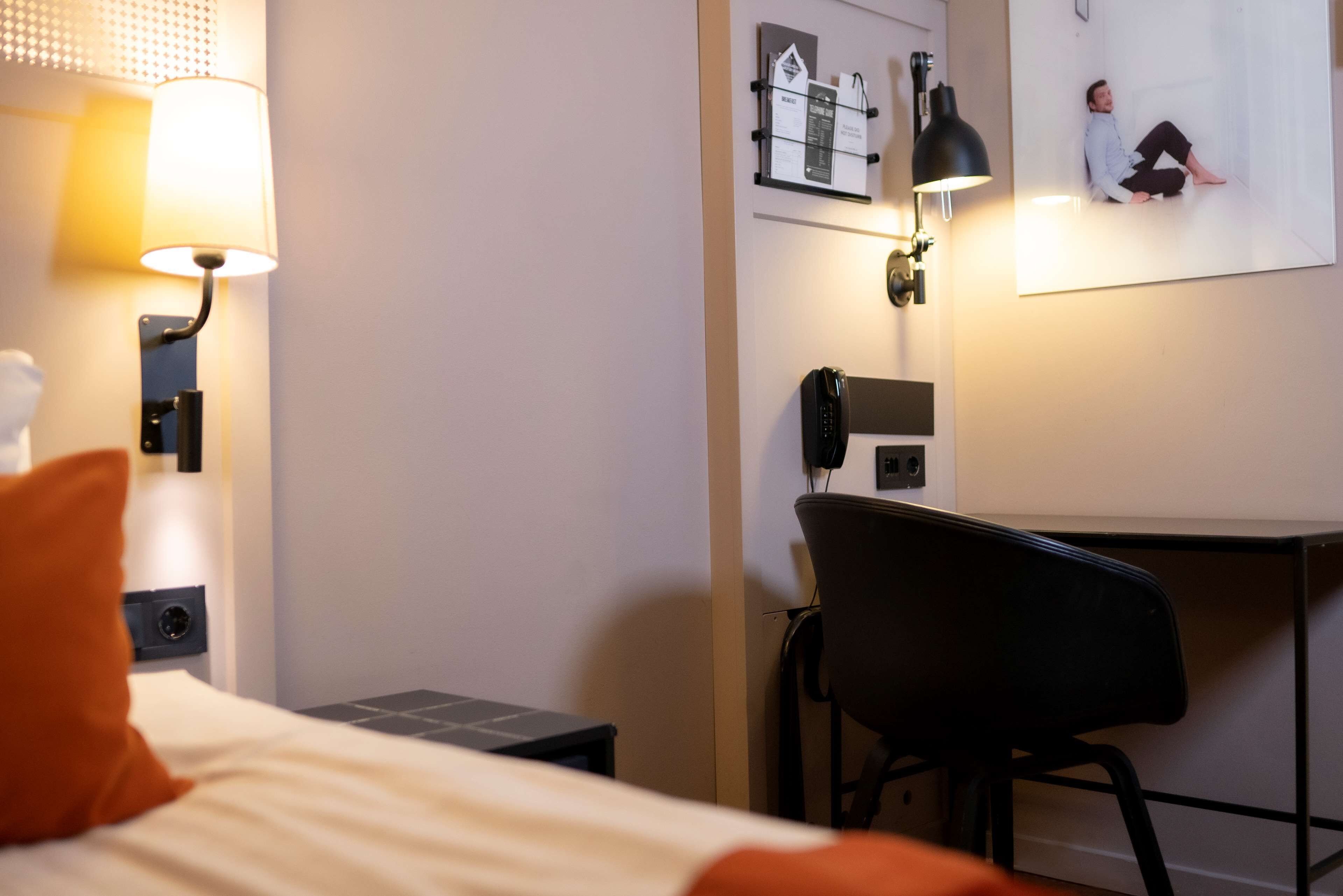 A hotel room with a bed, a desk, a chair, a lamp, and a wall-mounted phone.