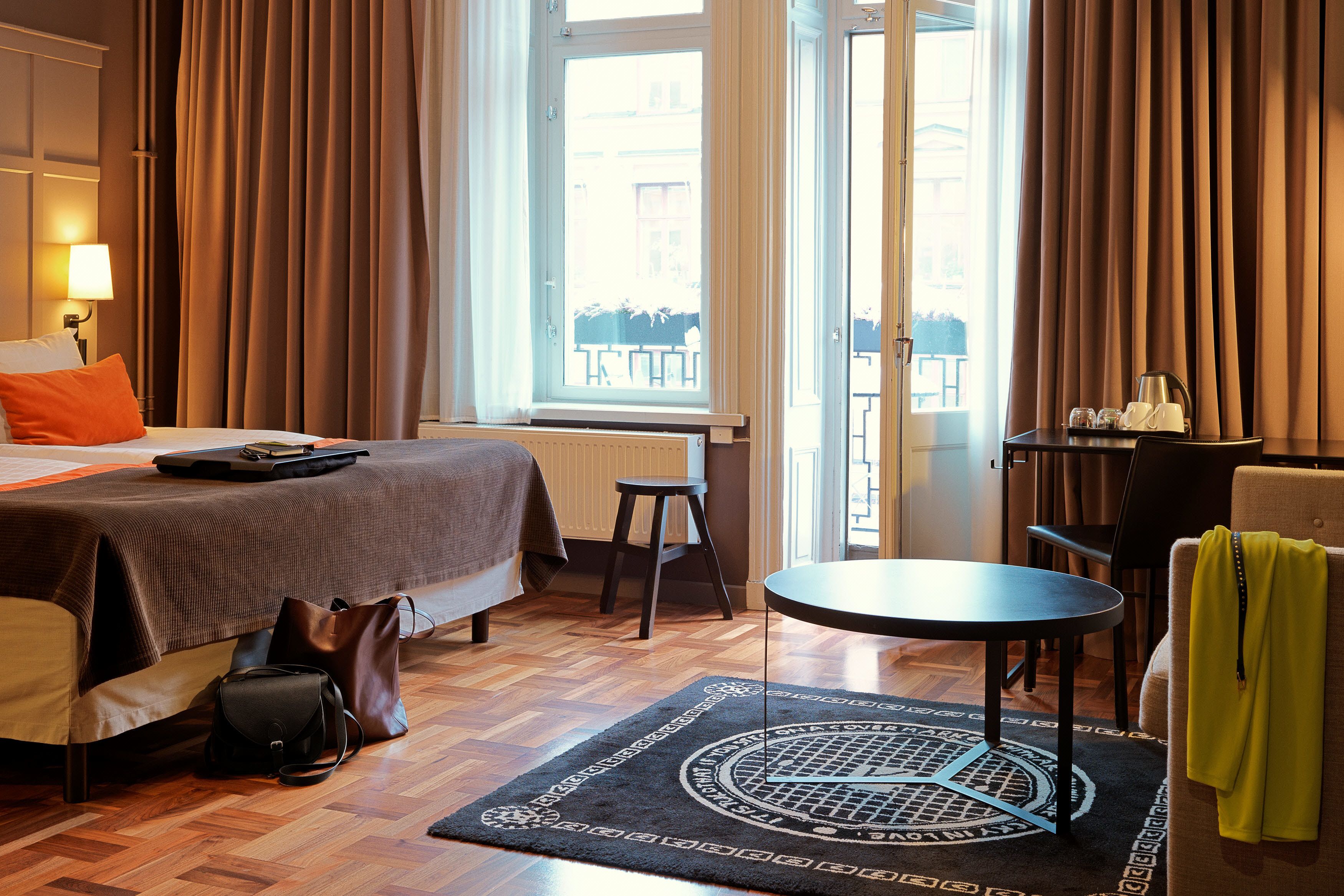 Habitación de hotel con cama, escritorio, silla, una mesita, un radiador y ventana con cortinas.