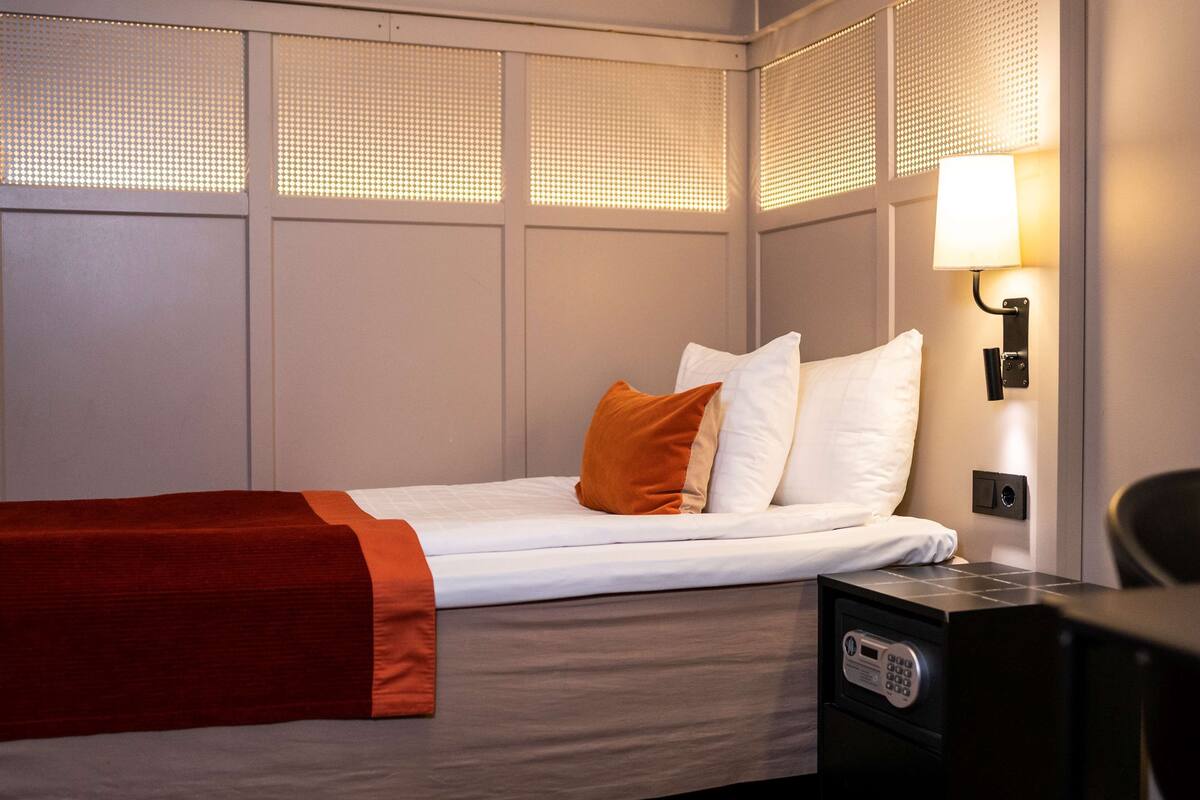 Una cama bien tendida con almohadas blancas y anaranjadas, una colcha roja y una mesita de noche negra con un reloj.