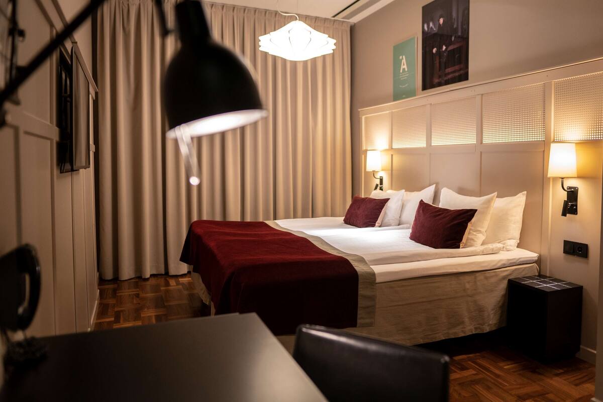 Habitación de hotel con cama, lámparas de noche, una manta roja, piso de madera y una pequeña mesa.