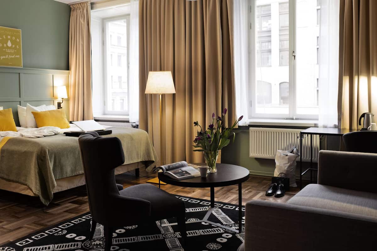 Habitación de hotel con cama, escritorio, silla, lámpara, un jarrón con flores y un radiador.