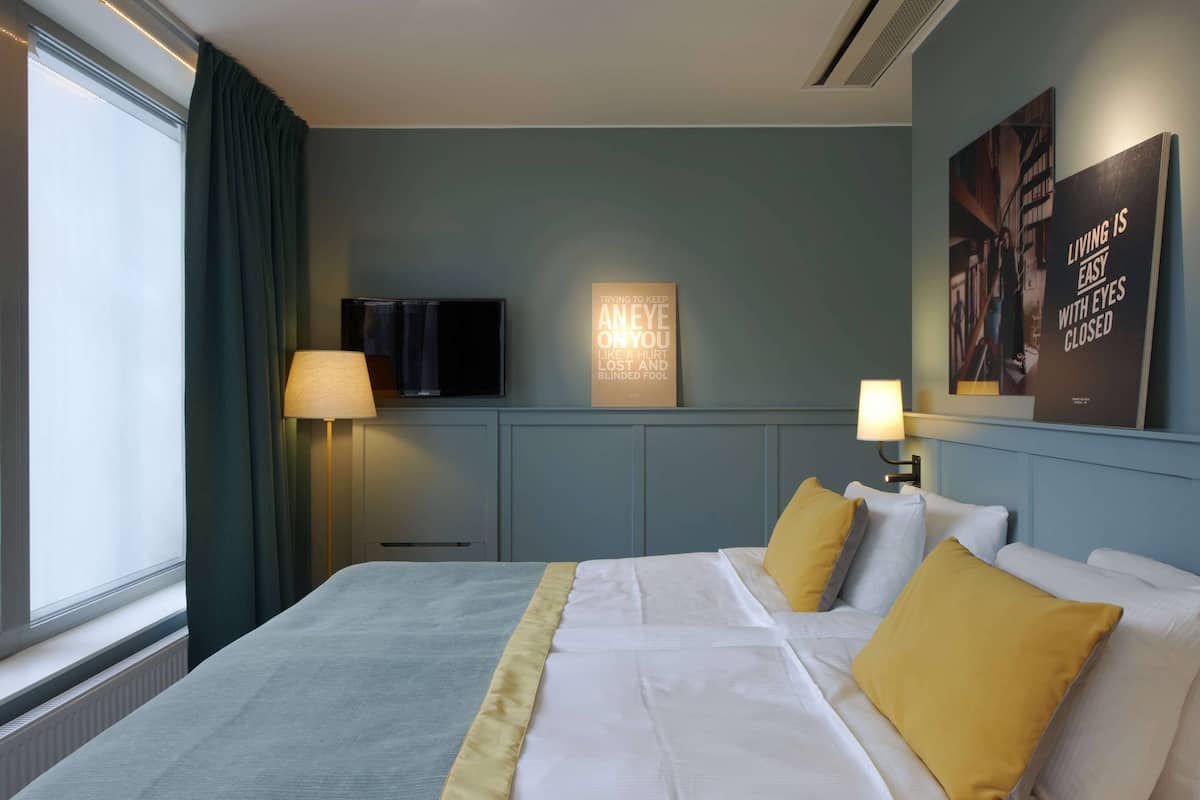 Habitación de hotel con una cama, dos lámparas de noche, un televisor de pantalla plana y dos carteles motivacionales en la pared.