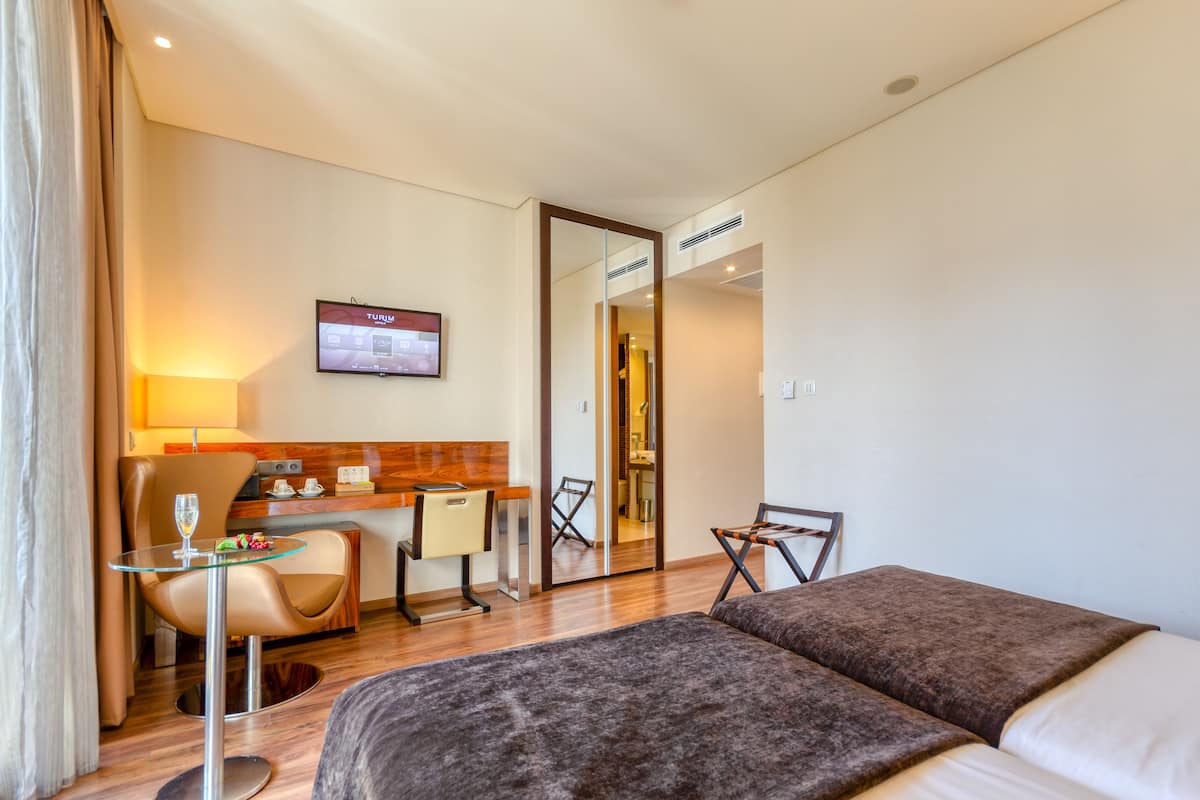 Ein Hotelzimmer mit einem Bett, einem Schreibtisch, einem Sessel, einem kleinen Tisch mit Glasplatte und einem an der Wand befestigten Fernseher.