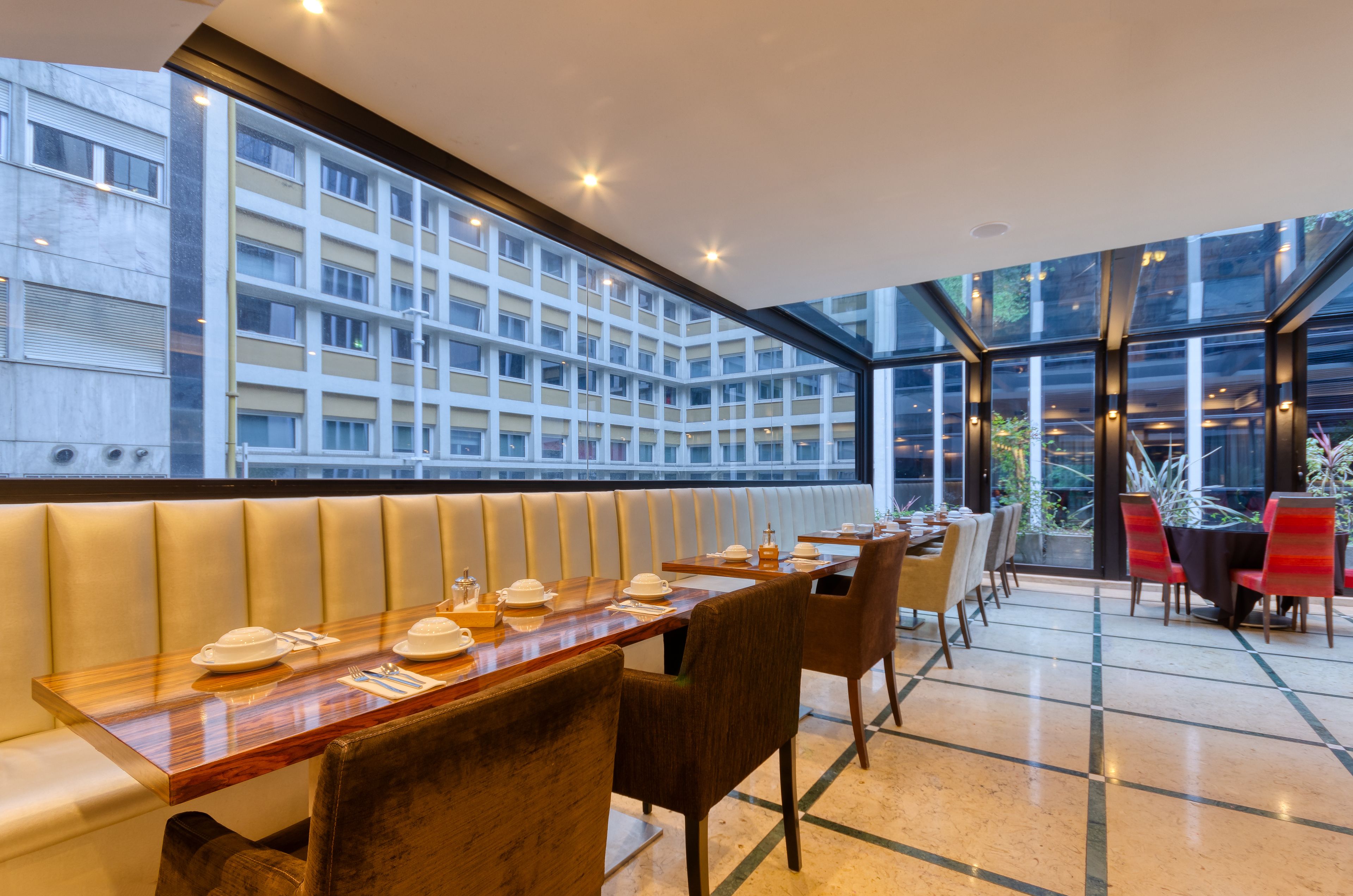 A restaurant with a long wooden table set for dining, a high ceiling, and large windows offering a view of a building across the street.