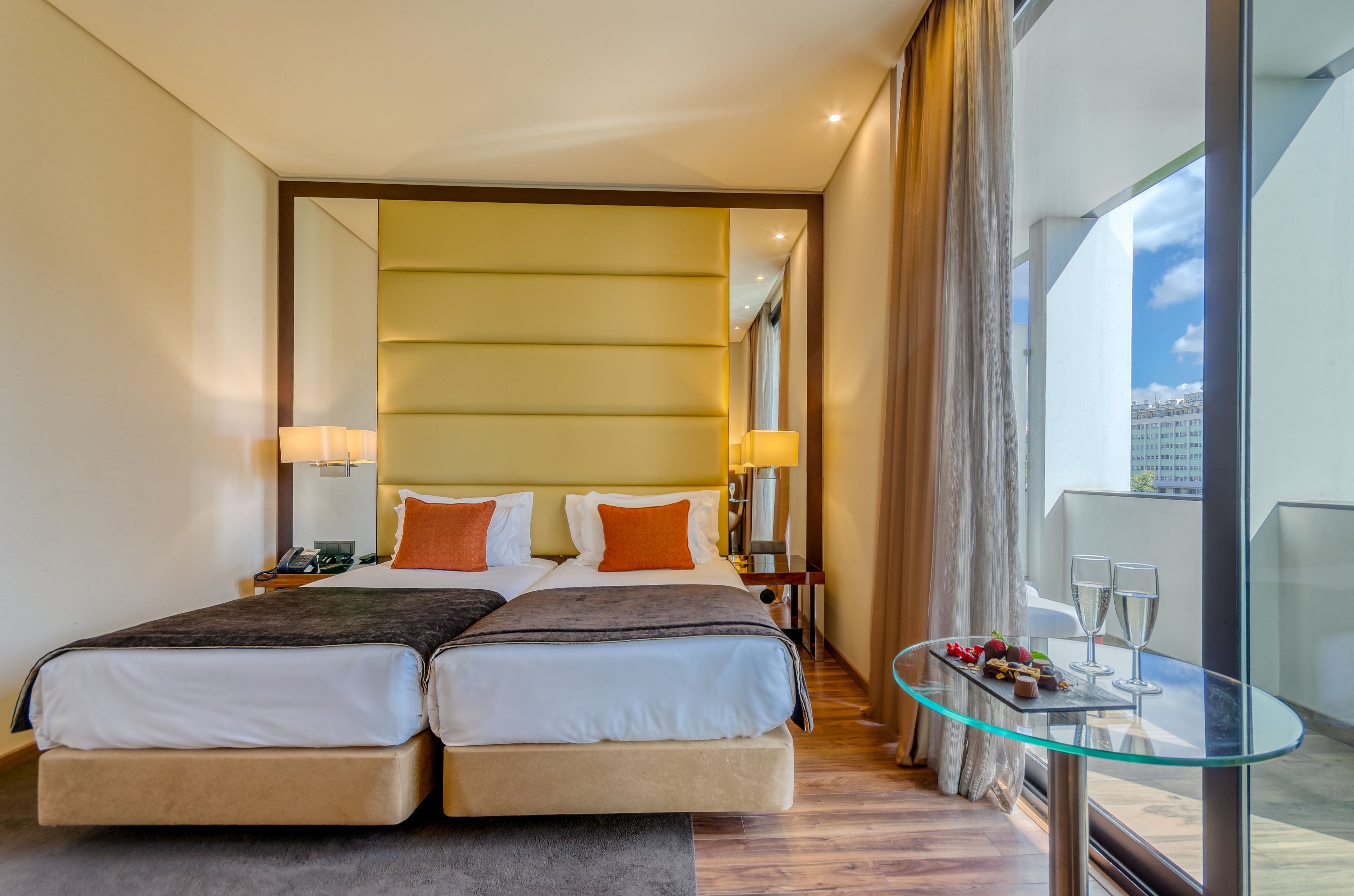 Une chambre d’hôtel avec un lit double, des tables de chevet, une table en verre avec deux verres, et une vue sur la ville grâce à de grandes fenêtres.