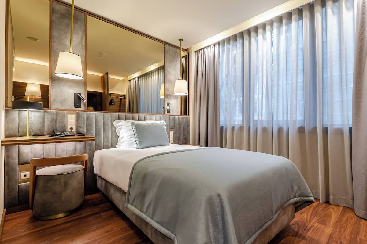 Ein modernes Hotelzimmer mit einem großen Bett, einem Nachttisch mit Lampe, einem Spiegel und Blick ins Freie durch transparente Vorhänge.