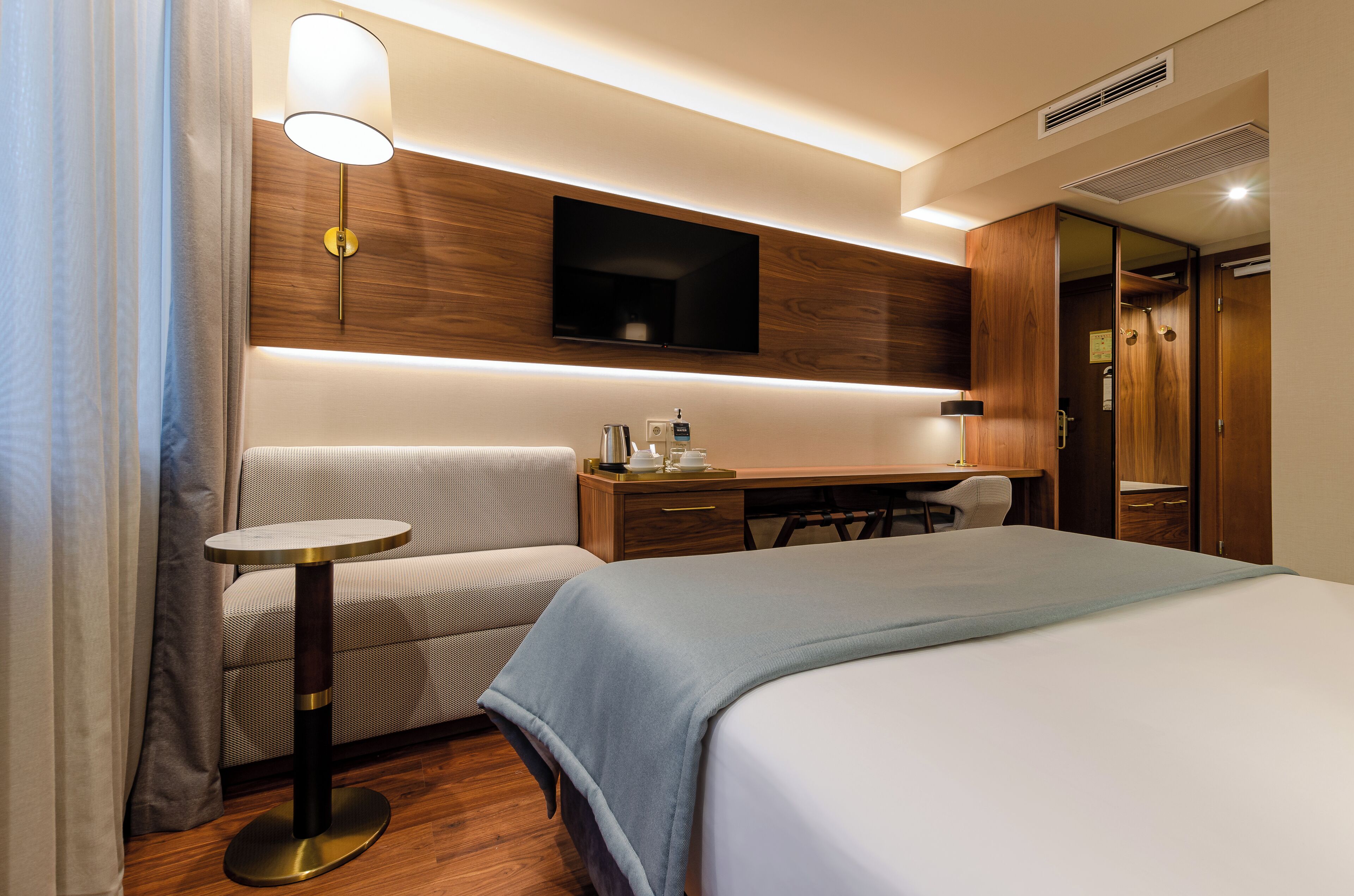 A hotel room with a bed, a desk, a chair, a small table, a TV mounted on the wall, and a wardrobe.