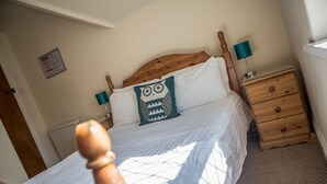 A bedroom with a wooden headboard, a bed with white bedding, a teal owl pillow, a wooden nightstand with a lamp, and a small wooden dresser. - St Breca (Newquay)