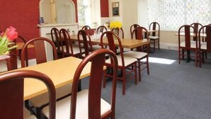A dining area with wooden tables and chairs, a red accent wall, and a window with a patterned curtain. - St Breca (Newquay)