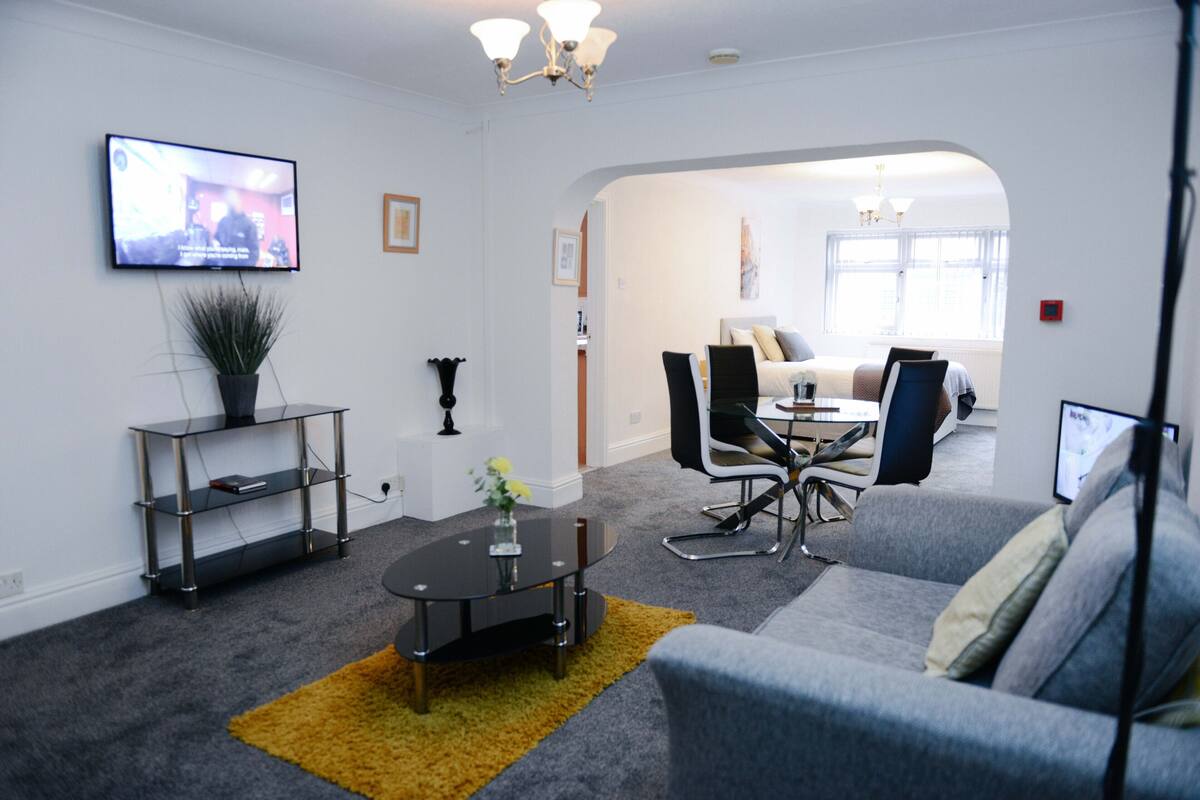 Ein modernes Wohnzimmer mit einem grauen Sofa, einem Couchtisch und einem an der Wand montierten Fernseher. Durch einen Bogen ist ein Essbereich mit Tisch und Stühlen zu sehen.