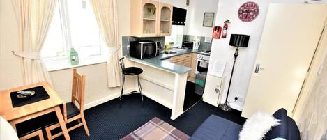 Una cocina compacta con microondas, lavamanos y armarios, un comedor con mesa y sillas, y una zona de descanso acogedora con cama y almohadas.