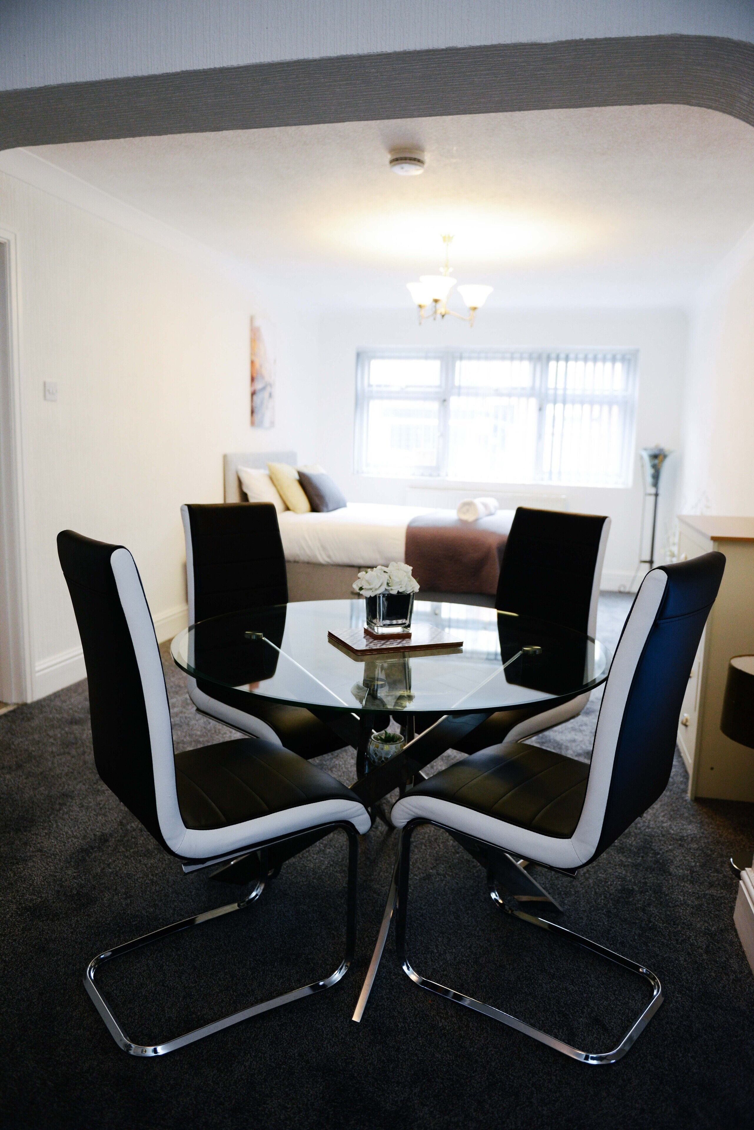 A modern dining area with a glass table and black and white chairs, a sofa in the background, and a chandelier.