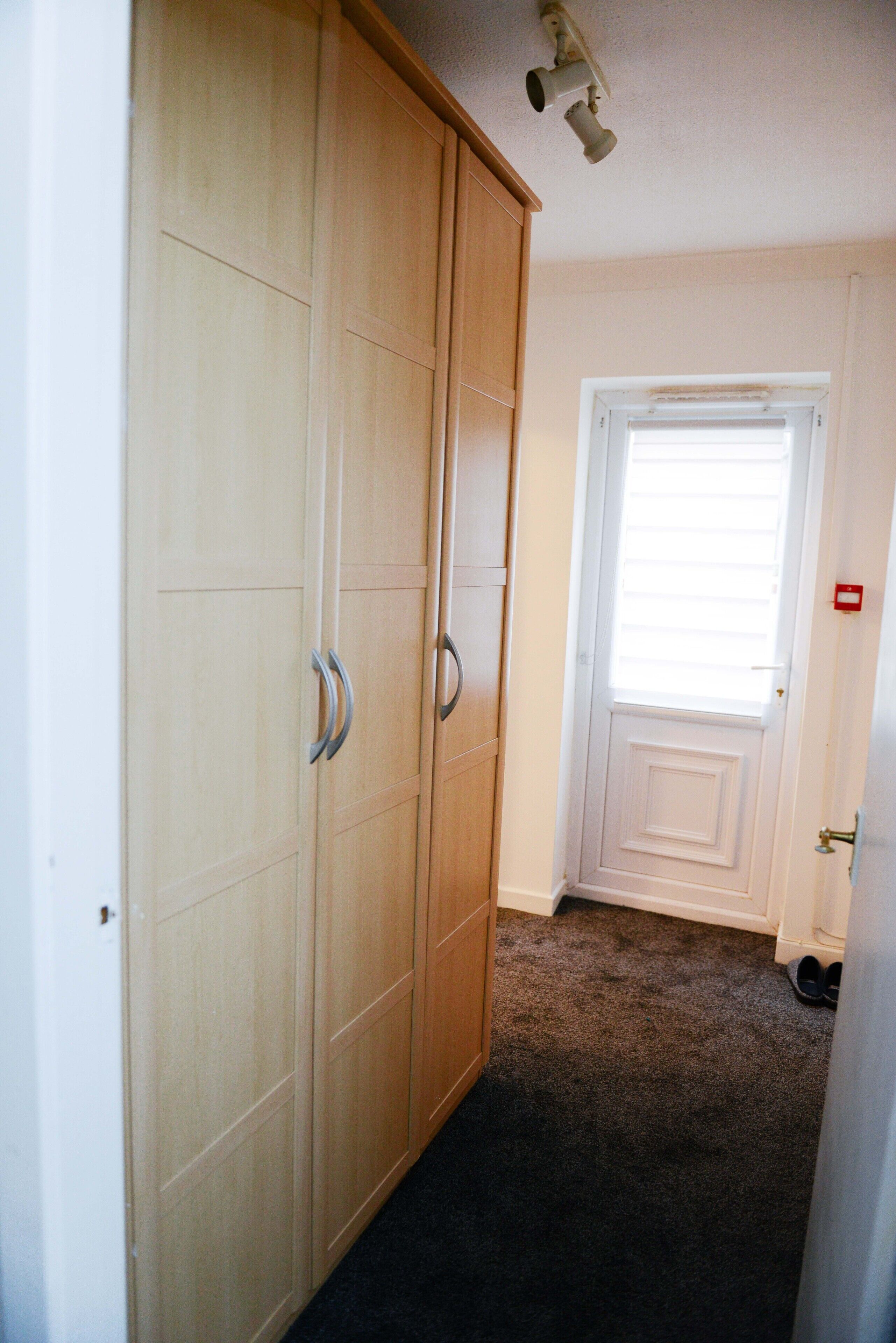 A room with a large wooden wardrobe, a white door with a window, and a carpeted floor.
