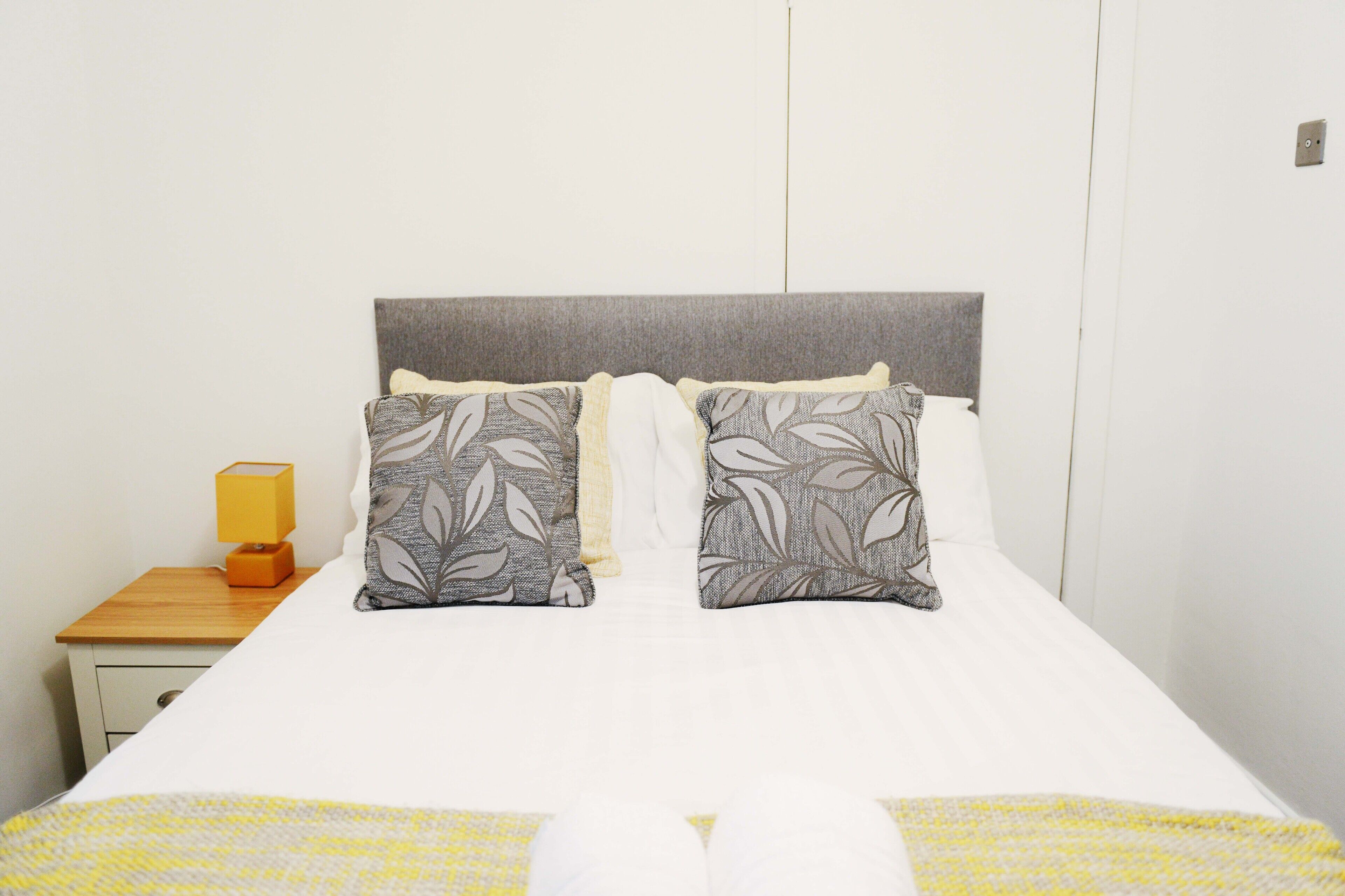 A neatly made bed with a white duvet and two pillows with a leaf pattern, a wooden nightstand with a lamp, and a white wardrobe in a hotel room.