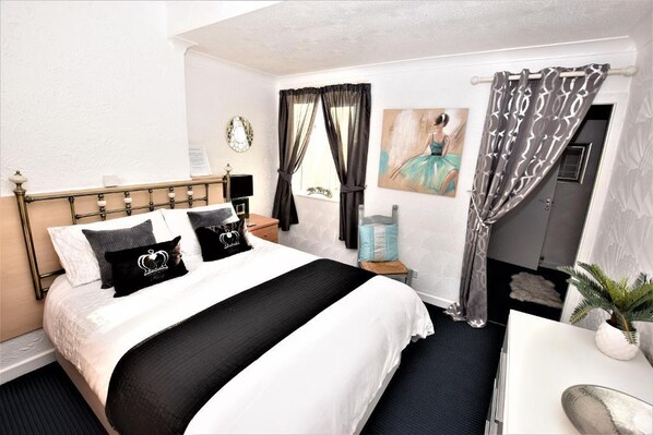 Habitación de hotel con cama, escritorio, silla, planta en maceta, cuadro y ventana con cortinas.