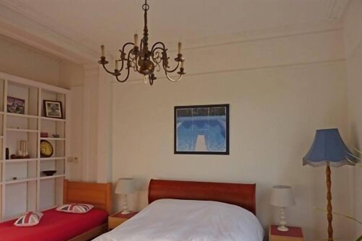 A hotel room with a bed, a bedside table, a nightstand, a lamp, a bookshelf, and a chandelier.