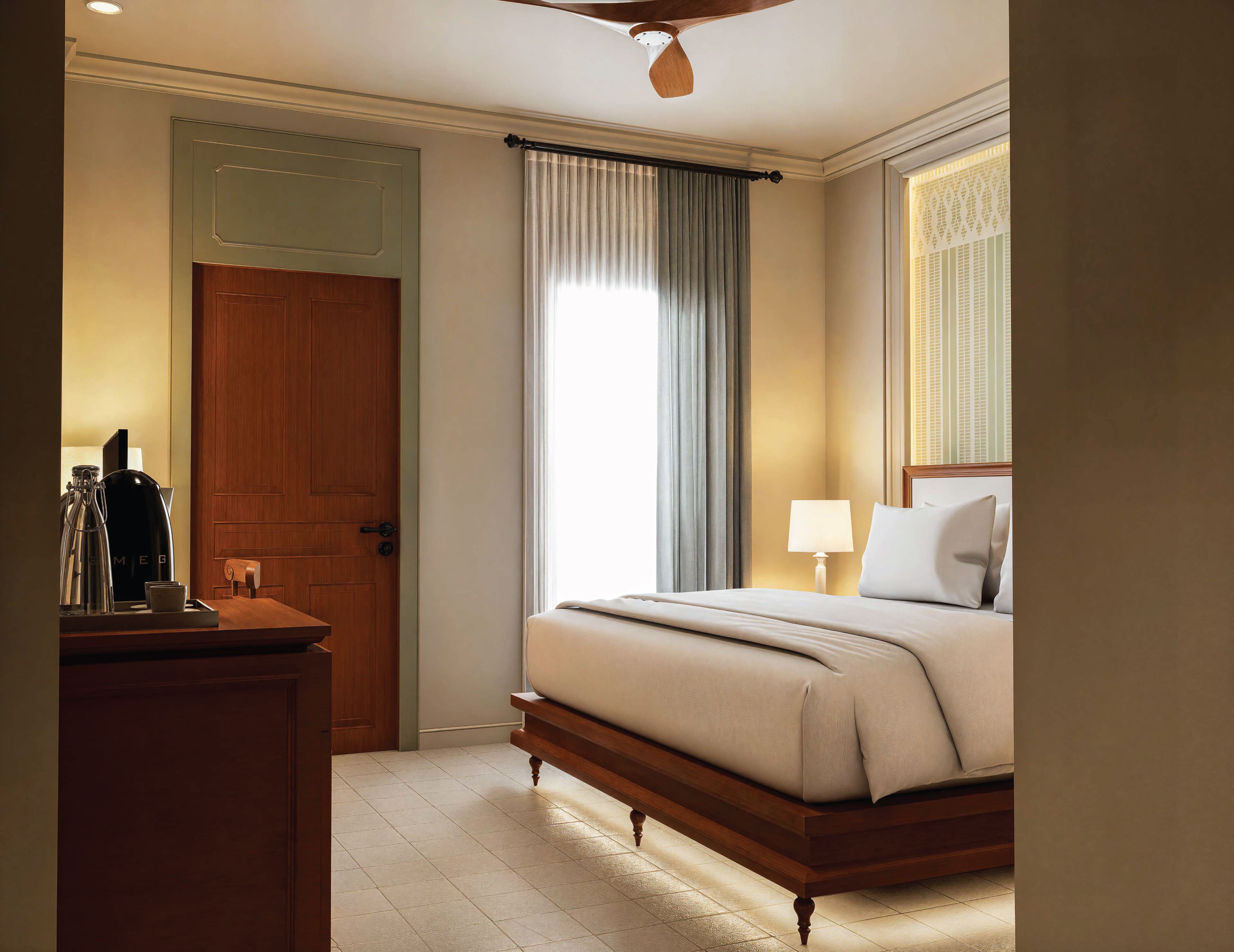 Habitación de hotel con una cama de madera, una mesita de noche con lámpara, una ventana con cortinas y una puerta.