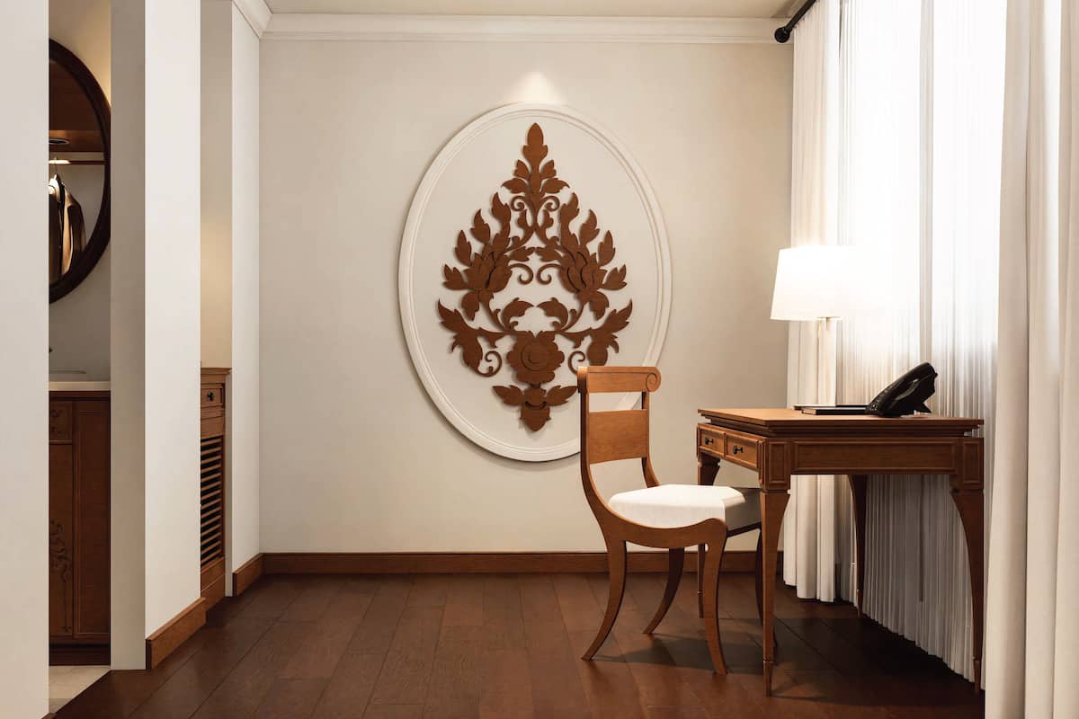 Quarto de hotel com uma cadeira de madeira, uma escrivaninha com abajur e um objeto decorativo na parede.