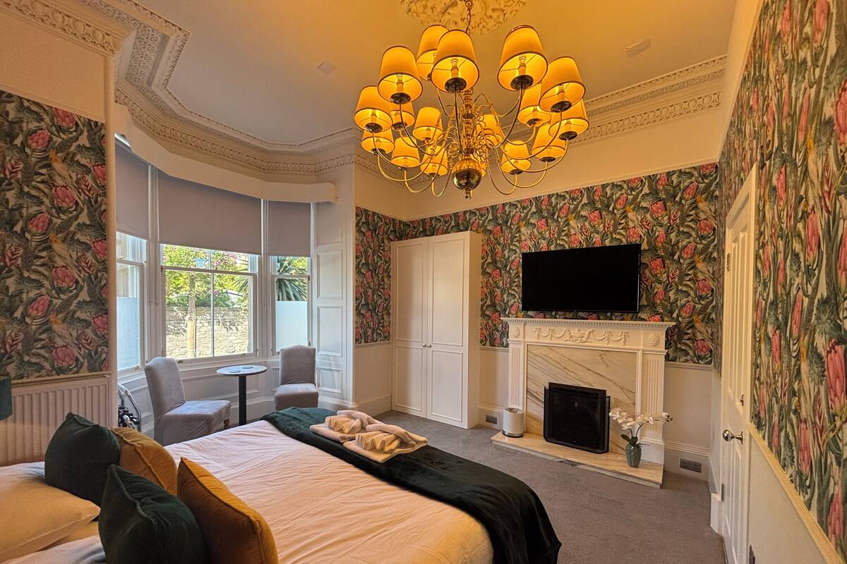 A bedroom with a large bed, a fireplace, a television, and a window with a view of greenery.
