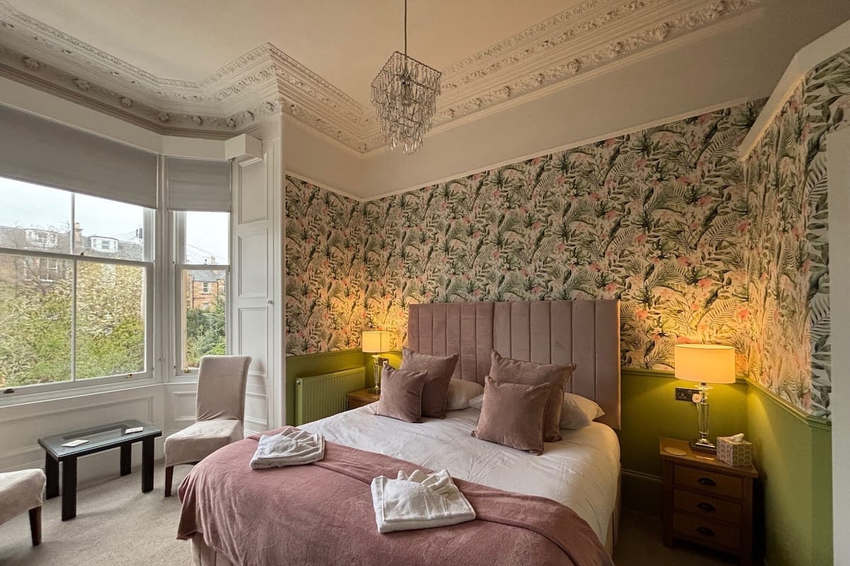 A bedroom with a large bed, two bedside tables, a chair, a window, and a patterned wallpaper.