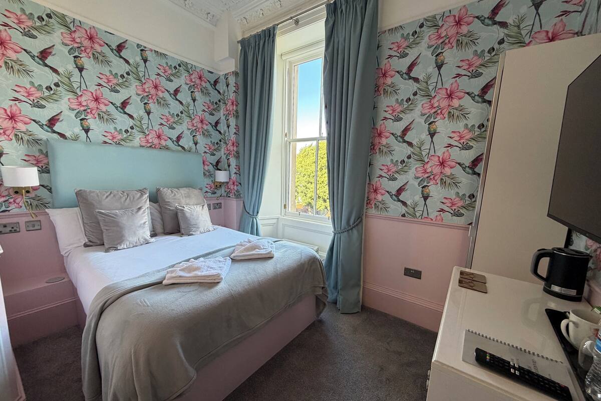 A bedroom with a bed, pillows, a nightstand, a television, a coffee maker, and a window with curtains.