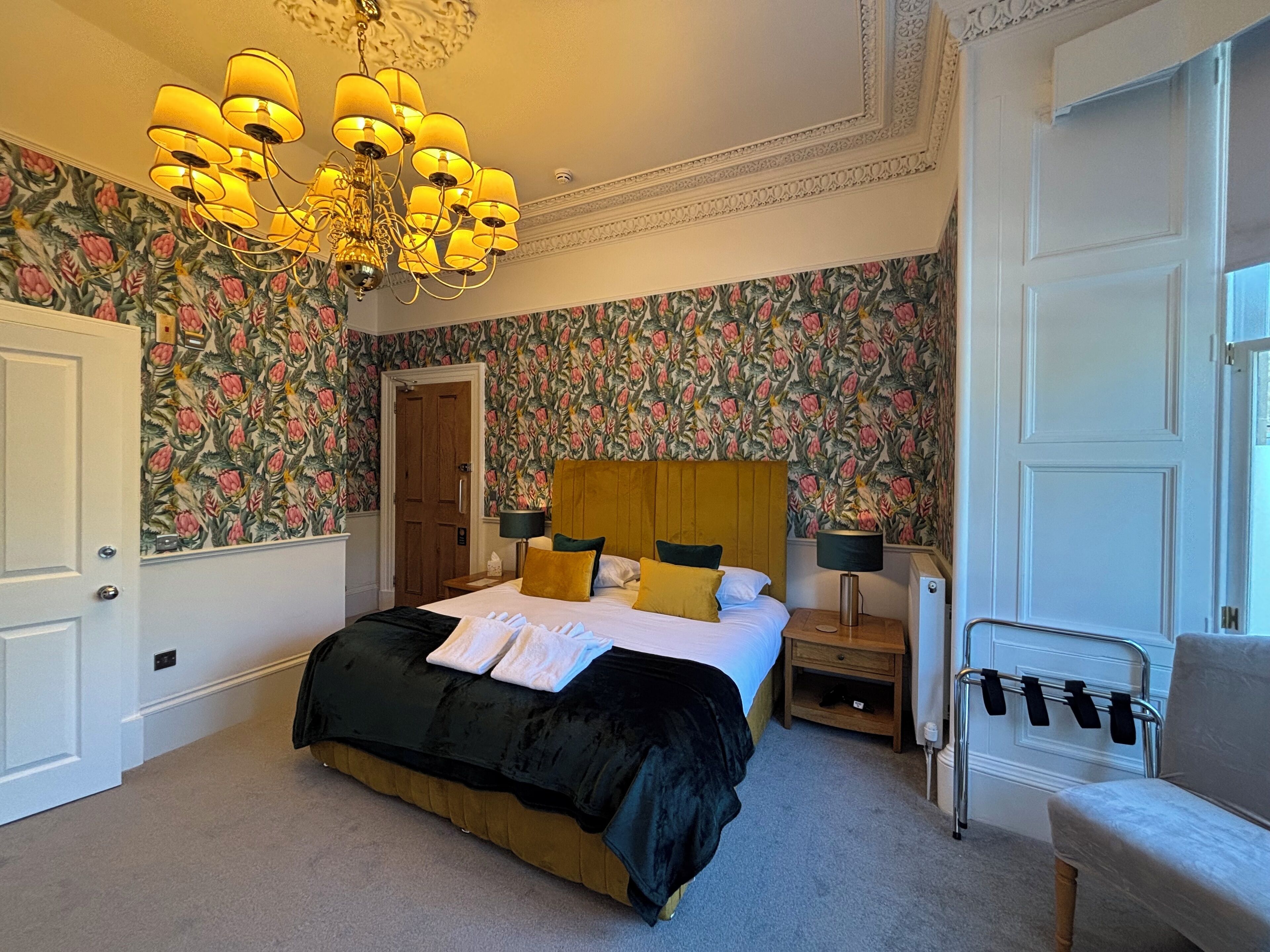 A bedroom with a floral wallpaper, a bed with a black and gold bedspread, a wooden headboard, two bedside tables with lamps, and a chair with a coat rack.