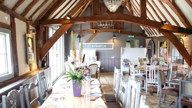 Una larga mesa de madera para comer, dispuesta con platos, cubiertos y copas, rodeada de sillas blancas en un interior rústico con vigas de madera a la vista y un candelabro.
