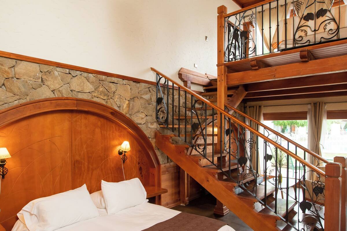 Een slaapkamer met een groot bed, houten hoofdeinde, stenen muur en een trap naar een hoger gelegen verdieping.