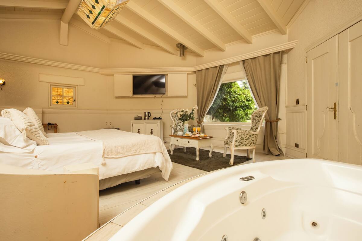 Een slaapkamer met een groot bed, een zithoek met een tafel en stoelen, een televisie en een raam met gordijnen.