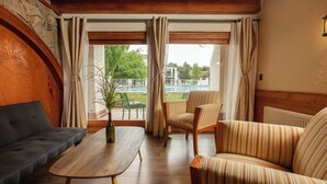 Una sala de estar acogedora con un sofá, un sillón y una mesa de centro. Un ventanal con cortinas ofrece vistas a una piscina y zonas verdes.