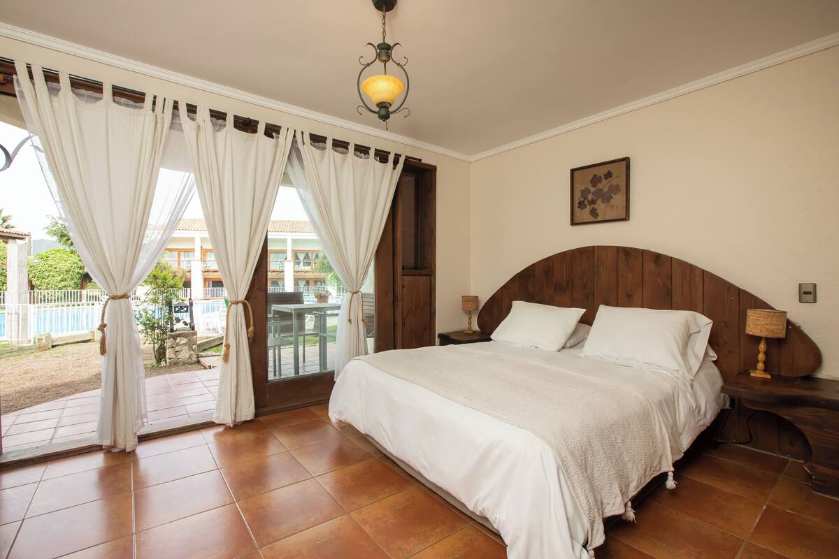Een slaapkamer met een groot bed, houten hoofdeinde en uitzicht op een zwembad en gebouwen.