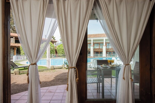 A view through a window with curtains, revealing a pool area with a table and chairs, and a building with a tiled roof in the background.