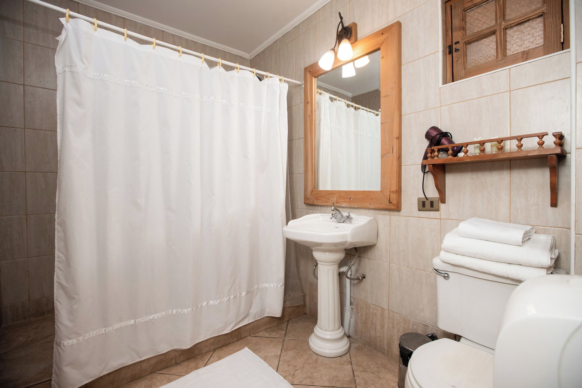 A bathroom with a shower curtain, a pedestal sink, a mirror, a toilet, and a shelf with towels.