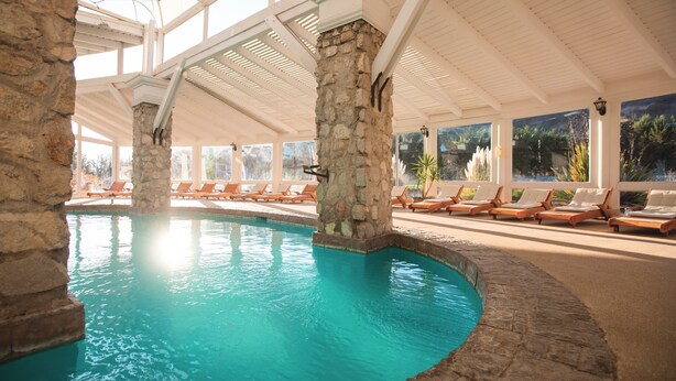 Una piscina interior con pilares de piedra, sillones de descanso y techo de cristal.