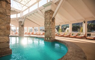 Una piscina interior con pilares de piedra, sillones de descanso y techo de cristal.