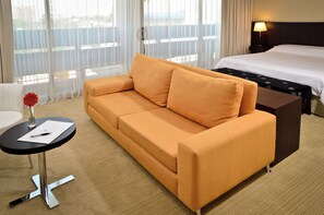 A hotel room with a sofa, a small table with a pen and paper, a lamp, and a bed. - Bagú Urbano (Posadas)
