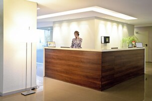 Un mostrador de recepción con una mujer de pie detrás, una pantalla digital y una planta en la encimera.