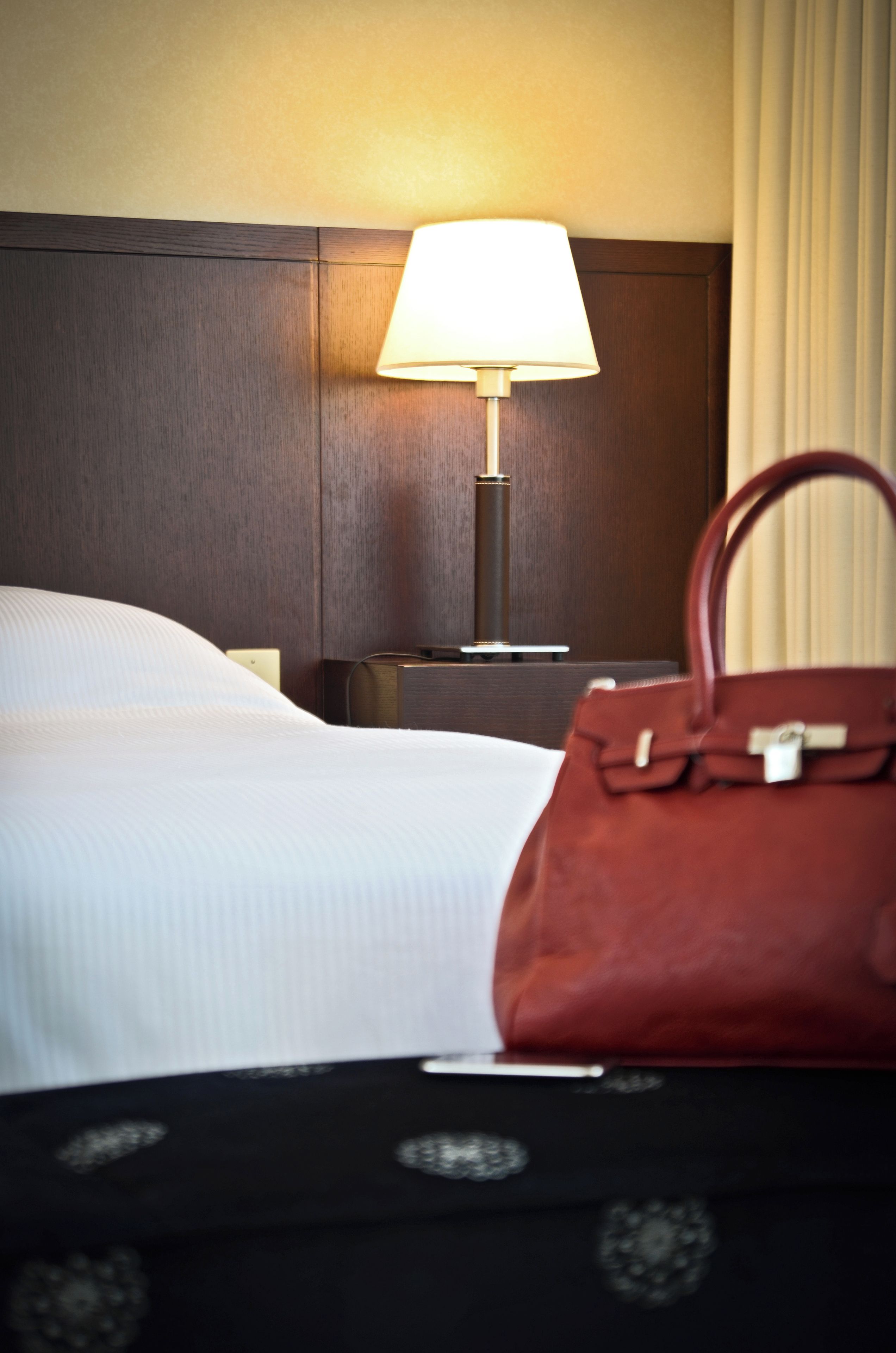 A hotel room with a bed, a nightstand, a lamp, and a handbag.
